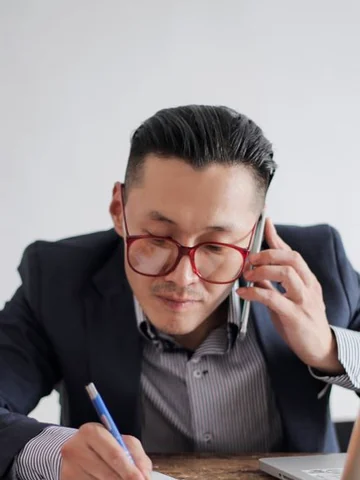 Manager in a navy suit and glasses on a call while taking notes at his desk